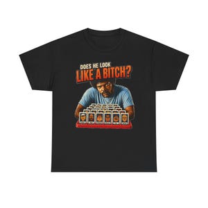 Puede incluir: Una camiseta negra de manga corta con un estampado gráfico de un hombre con camisa azul jugando a un juego de mesa con pequeñas cartas de personajes. Sobre la figura, el texto dice DOES HE LOOK LIKE A BITCH? en letras naranjas y blancas.