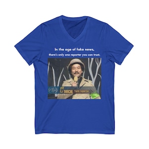 May include: Royal blue V-neck t-shirt featuring a graphic of a man holding a microphone, with the text "In the age of fake news, there's only one reporter you can trust." The graphic also includes the text "guy LE DOUCHE field reporter."