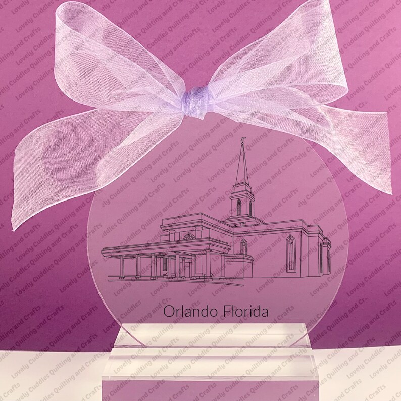 May include: A clear glass ornament with a black line drawing of a church building. The ornament is on a white base and has a pink background. The text "Orlando Florida" is printed on the ornament.