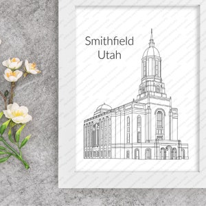 May include: A framed black and white line drawing of the Smithfield Utah temple. The artwork is in a white frame and features the name of the city and state. The image is on a gray surface with white flowers.