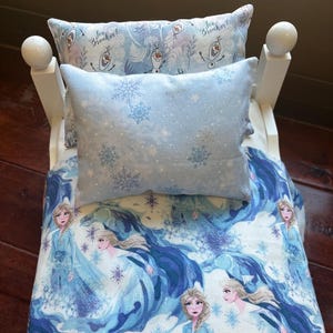 May include: A doll bed with bedding featuring a light blue snowflake pattern and images of a princess. The set includes a pillow and a comforter. The headboard and footboard are white.
