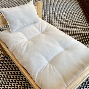 May include: A small, light-colored wooden pet bed with a white cushion and pillow. The bed frame is made of natural wood, and the cushion has a tufted design. The pillow is square and rests on the bed. The bed is designed for small pets.