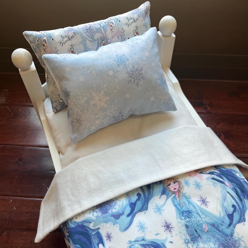 May include: A white wooden doll bed with two pillows and a blanket. The pillows are light blue with snowflake and character designs. The blanket and bedspread feature a blue and white Frozen theme.