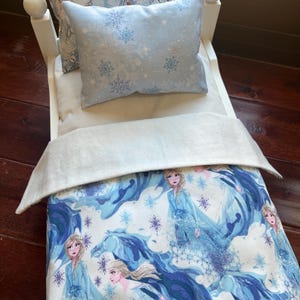 May include: A white doll bed with a blue and white comforter and pillows. The comforter features a print of a woman in a blue dress with a blue and white background. The pillows have snowflake and character prints.