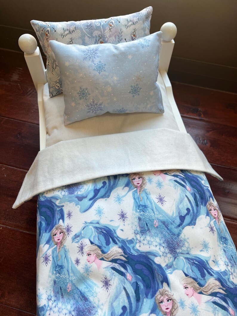 May include: A white doll bed with a blue and white comforter and pillows. The comforter features a print of a woman in a blue dress with a blue and white background. The pillows have snowflake and character prints.
