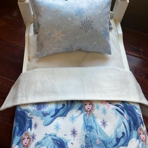May include: A miniature white bed with bedding featuring a winter theme. The bedding includes a light blue pillow with snowflake patterns, a white blanket, and a comforter with a blue and white illustration of a character from a popular animated film.
