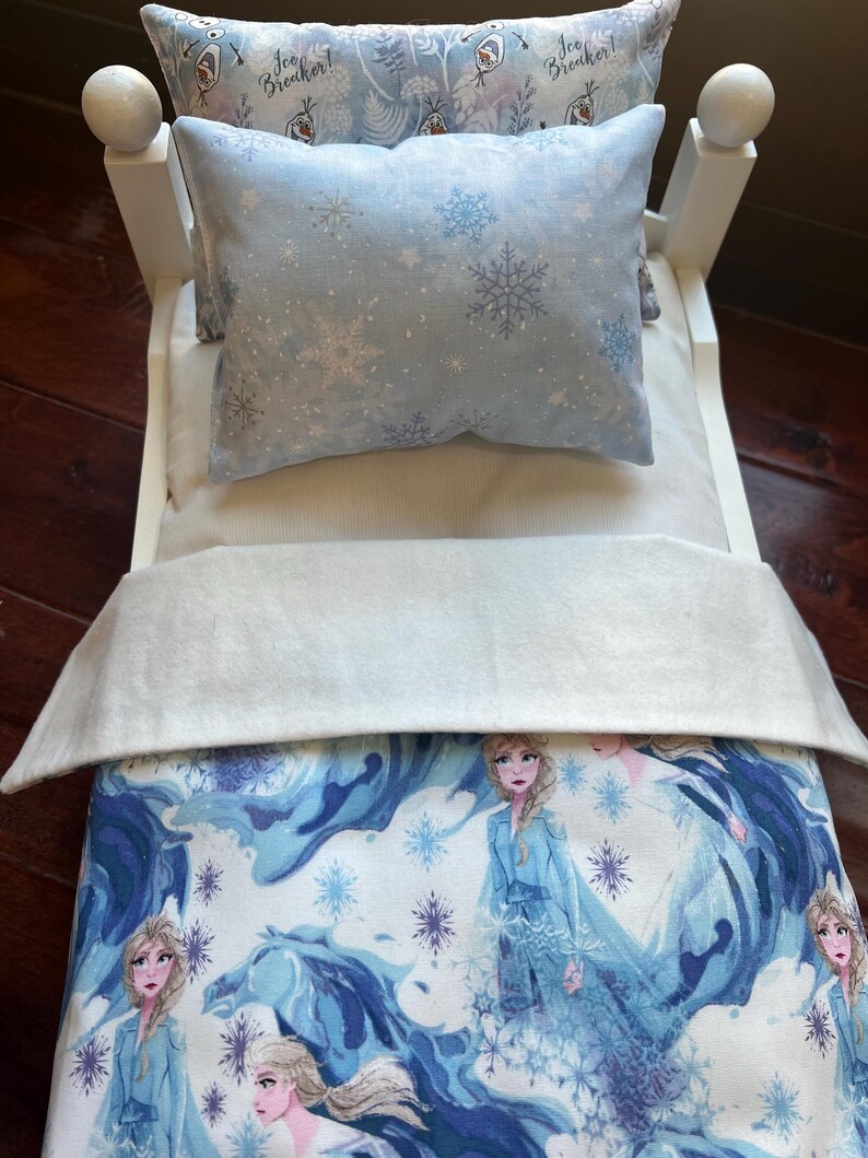 May include: A miniature white bed with bedding featuring a winter theme. The bedding includes a light blue pillow with snowflake patterns, a white blanket, and a comforter with a blue and white illustration of a character from a popular animated film.