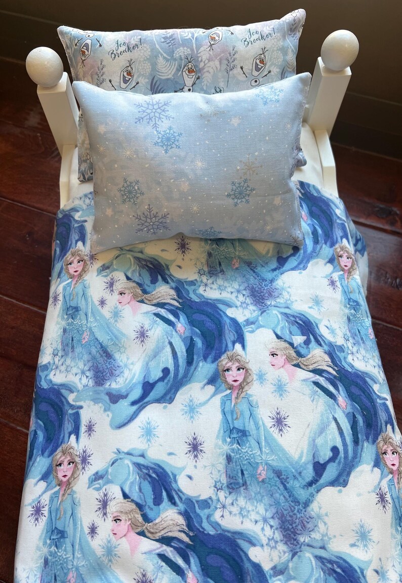 May include: A doll bed with bedding featuring a light blue snowflake pattern and images of a princess. The set includes a pillow and a comforter. The headboard and footboard are white.