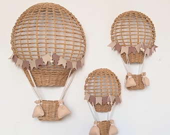 Op de afbeelding: Drie wanddecoraties in de vorm van heteluchtballonnen, gemaakt van gevlochten lichtbruin riet. Elke ballon heeft een ronde bovenkant, een klein mandje opgehangen aan witte koorden en een slinger van kleine beige en mauve stoffen vlaggetjes.