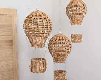 Op de afbeelding: Drie hangende heteluchtballon-decoraties gemaakt van lichtbruin gevlochten riet. Elke ballon heeft een rond bovengedeelte dat met witte koorden is verbonden aan een kleine cilindervormige mand, versierd met kleine houten kralen aan de bovenkant.