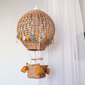 Könnte beinhalten: Eine hängende Leuchte in Form eines Heißluftballons aus geflochtenem hellbraunem Rattan. Sie besteht aus einem runden Ballonabschnitt, einem kleinen quadratischen Korb und einer dekorativen Wimpelkette in Senfgelb, Blau und Beige.
