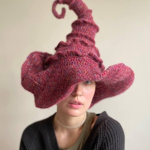 K&ouml;nnte beinhalten: Ein burgunderfarbener, handgestrickter Hexenhut mit einer gekr&auml;uselten Spitze und breiter Krempe. Der Hut ist aus einem strukturierten Garn mit Farbtupfern gefertigt, was ihm ein rustikales Aussehen verleiht. Das Design des Hutes ist skurril und einzigartig.