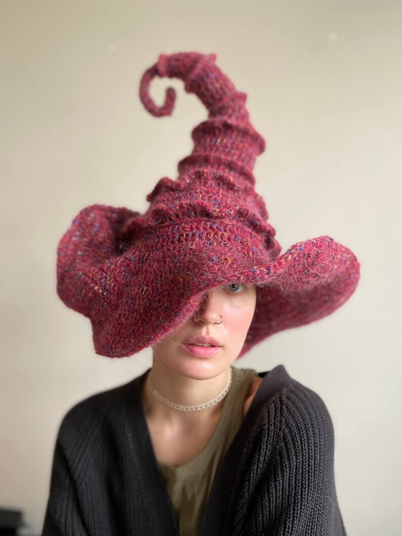 K&ouml;nnte beinhalten: Ein burgunderfarbener, handgestrickter Hexenhut mit einer gekr&auml;uselten Spitze und breiter Krempe. Der Hut ist aus einem strukturierten Garn mit Farbtupfern gefertigt, was ihm ein rustikales Aussehen verleiht. Das Design des Hutes ist skurril und einzigartig.