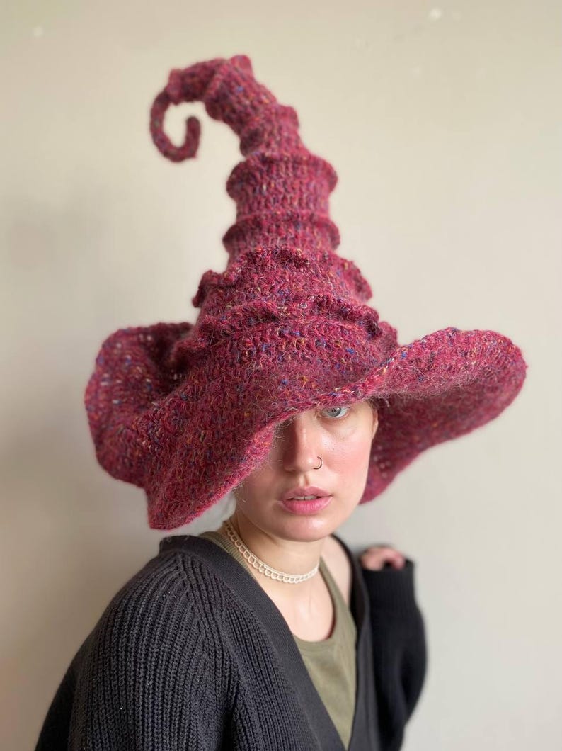 K&ouml;nnte beinhalten: Ein handgestrickter Hexenhut in Burgunder- und Lilat&ouml;nen. Der Hut hat eine breite, flatternde Krempe und eine hohe, spiralf&ouml;rmige Spitze. Das strukturierte Garn verleiht diesem einzigartigen Accessoire Tiefe und visuelles Interesse.