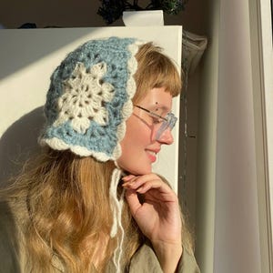 May include: A handmade crochet bonnet in light blue and cream. The bonnet features a floral design in the center and a scalloped edge. White ties hang from the sides. The bonnet is a stylish accessory.