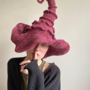 K&ouml;nnte beinhalten: Handgestrickter Hexenhut in leuchtendem Magenta mit breiter Krempe und hohem, spiralf&ouml;rmigem Zipfel. Das strukturierte Strickmuster verleiht diesem einzigartigen Accessoire Tiefe und visuelles Interesse.