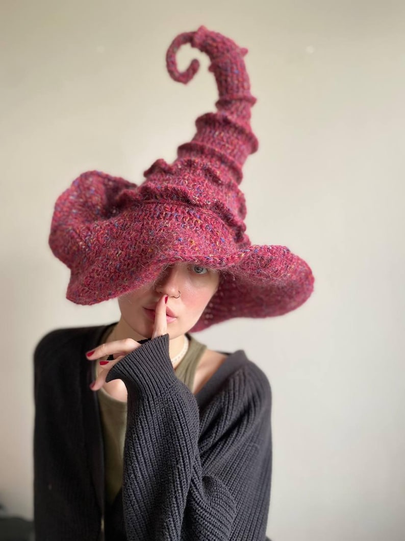 K&ouml;nnte beinhalten: Handgestrickter Hexenhut in leuchtendem Magenta mit breiter Krempe und hohem, spiralf&ouml;rmigem Zipfel. Das strukturierte Strickmuster verleiht diesem einzigartigen Accessoire Tiefe und visuelles Interesse.