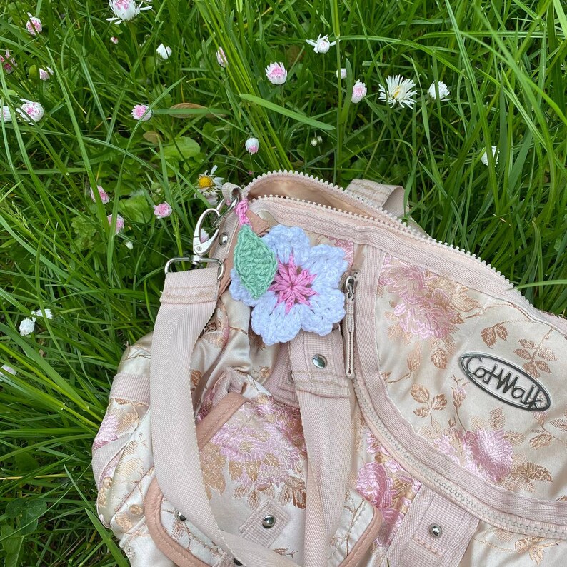 May include: A pink and white crocheted flower with a green leaf attached to a light pink handbag with a floral pattern. The bag has a zipper and a strap. The text "CatWalk" is visible on the bag.