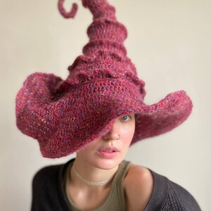 K&ouml;nnte beinhalten: Ein handgestrickter Hexenhut in Burgunderrot mit einer hohen, spiralf&ouml;rmigen Spitze und einer breiten, schlaffen Krempe. Der Hut ist aus einem strukturierten Garn mit Farbtupfern gefertigt. Ein einzigartiges Accessoire f&uuml;r Kost&uuml;m oder Alltag.