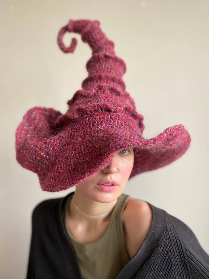 K&ouml;nnte beinhalten: Ein handgestrickter Hexenhut in Burgunderrot mit einer hohen, spiralf&ouml;rmigen Spitze und einer breiten, schlaffen Krempe. Der Hut ist aus einem strukturierten Garn mit Farbtupfern gefertigt. Ein einzigartiges Accessoire f&uuml;r Kost&uuml;m oder Alltag.
