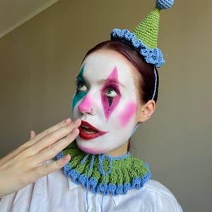 May include: A handmade crochet clown hat and collar set. The hat is green with a blue trim and a blue ball on top. The collar is green with a blue trim and blue ties. The person is wearing white face paint with pink, green, and red accents.