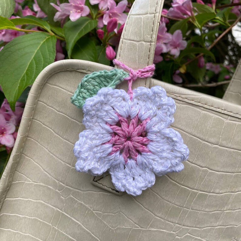 May include: A white crocheted flower with a pink center and green leaves attached to a light brown faux leather bag.