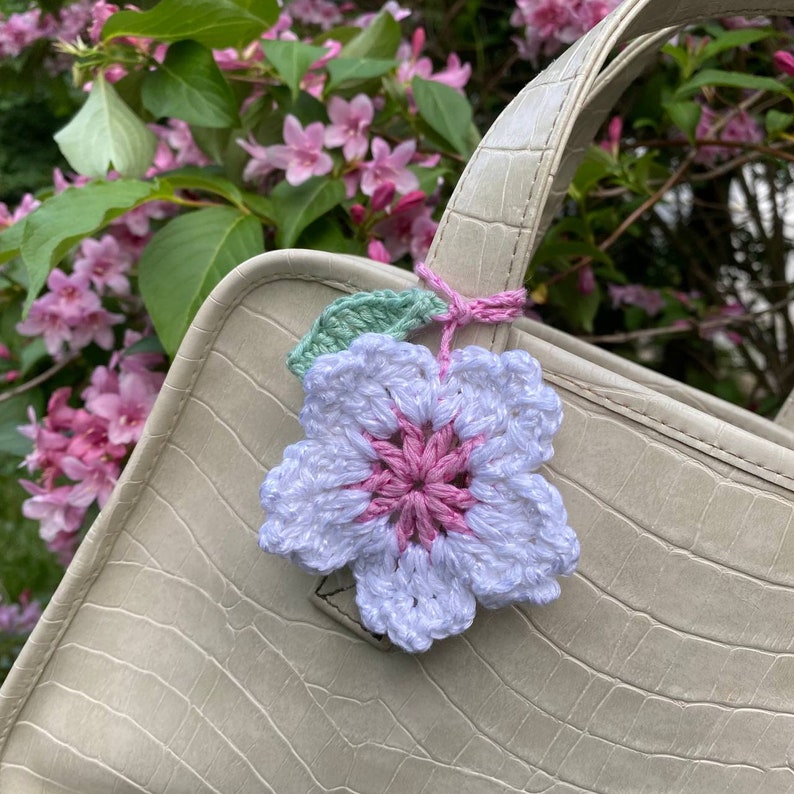 May include: A white crocheted flower with a pink center and green leaves attached to a beige handbag.