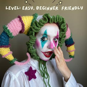 Puede incluir: Un sombrero de bufón de ganchillo multicolor con detalles en rosa, amarillo, verde y azul. El sombrero se usa con maquillaje de payaso y una camisa blanca. El texto "LEVEL: EASY, BEGINNER FRIENDLY" está encima del sombrero.