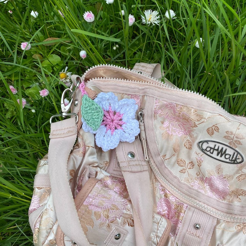 May include: A pink and white crocheted flower charm attached to a light pink bag with the word "CatWalk" printed on it.
