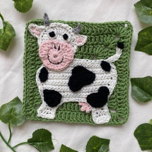 May include: A crocheted square featuring a white cow with black spots on a green background. The cow has a pink nose and a happy expression.
