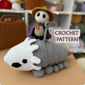 Puede incluir: Un armadillo de ganchillo con una pequeña figura montada en su espalda. El armadillo es gris y blanco, con ojos negros y una cola rosa. La figura lleva un sombrero amarillo y una capa morada. El texto "CROCHET PATTERN" es visible.