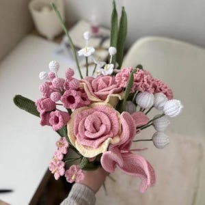 Puede incluir: Un ramo de flores de ganchillo en tonos de rosa y blanco. El ramo incluye rosas, lirios y otras flores delicadas.