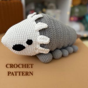 Puede incluir: Un juguete de peluche tejido a ganchillo con forma de criatura estilizada, con una cabeza blanca con elementos decorativos y un cuerpo gris. El juguete tiene ojos negros y pequeñas patas grises. El texto "CROCHET PATTERN" se muestra debajo.