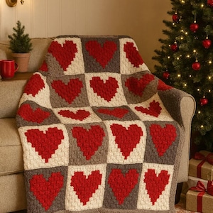 May include: A handmade crochet blanket with a repeating heart pattern. The blanket features red hearts on a white and grey background. The blanket is draped over a beige sofa. A Christmas tree is in the background.