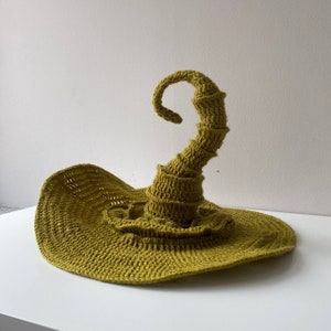 May include: A handmade, olive green crochet witch hat with a wide brim and a tall, spiraling point. The hat is made of a textured yarn, and the brim is slightly curved. The hat is displayed on a white surface.
