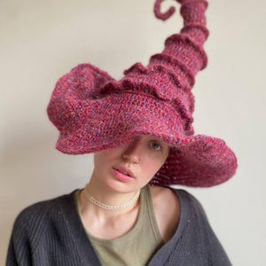 K&ouml;nnte beinhalten: Ein handgestrickter Hexenhut in leuchtendem Magenta. Der Hut hat eine breite, flatternde Krempe und eine hohe, spiralf&ouml;rmige Spitze. Das strukturierte Strickmuster zeigt Farbtupfer, die dem Design Tiefe verleihen. Ein einzigartiges Accessoire f&uuml;r Kost&uuml;m oder Alltag.