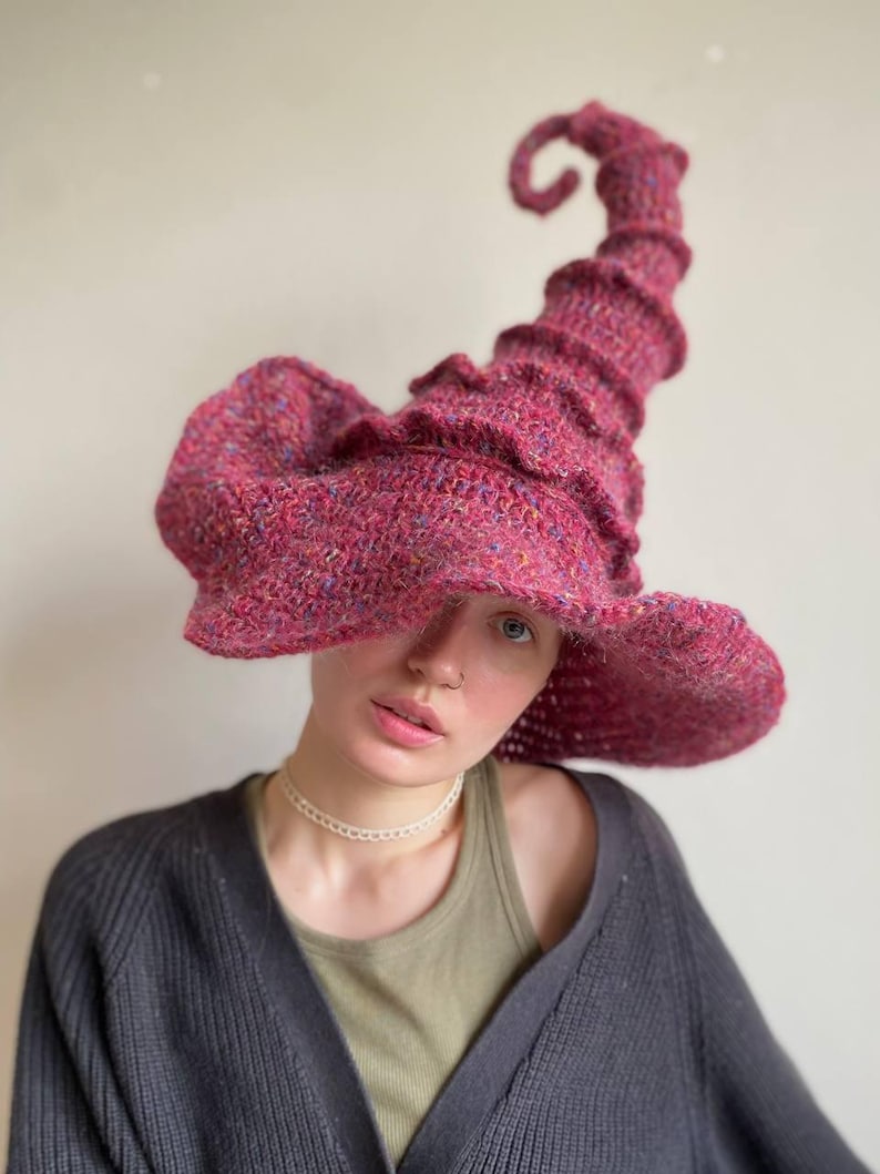 K&ouml;nnte beinhalten: Ein handgestrickter Hexenhut in leuchtendem Magenta. Der Hut hat eine breite, flatternde Krempe und eine hohe, spiralf&ouml;rmige Spitze. Das strukturierte Strickmuster zeigt Farbtupfer, die dem Design Tiefe verleihen. Ein einzigartiges Accessoire f&uuml;r Kost&uuml;m oder Alltag.