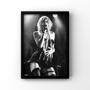 May include: Black and white photograph of a woman singing into a microphone on stage. She is wearing a black corset and fishnet stockings. The stage lights are shining on her.