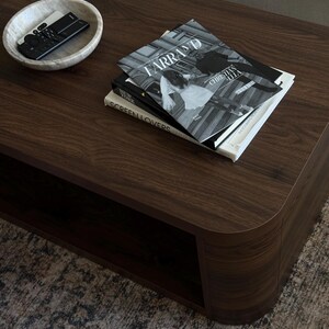 May include: A dark brown wooden coffee table with rounded edges. On the table are several books, including one titled "FARRAND" and a marble bowl holding a remote control. The table sits on a patterned rug.