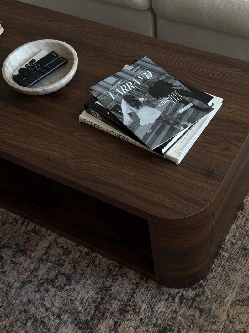 May include: A dark brown wooden coffee table with rounded edges. On the table are several books, including one titled "FARRAND" and a marble bowl holding a remote control. The table sits on a patterned rug.