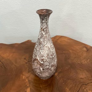 May include: A tall, slender ceramic vase featuring a flared rim and a tapered neck. The surface displays a textured, mottled glaze with a complex pattern of white, cream, and deep brown tones, creating a crackled, organic appearance throughout.