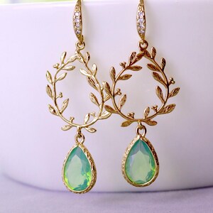 May include: Gold-toned earrings featuring a leaf-like design. Each earring has a teardrop-shaped, light green gemstone pendant. The earrings are hanging against a white background, with a blurred green and white backdrop.
