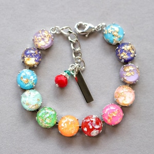 May include: A colorful bracelet featuring round beads in various colors, including turquoise, purple, pink, and green, with gold flecks. The bracelet has a silver chain and clasp, and a small charm with red and green accents.