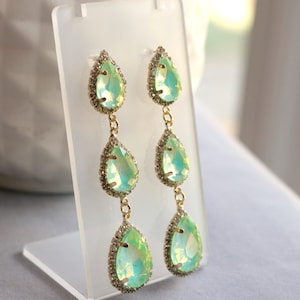 May include: Elegant, long drop earrings featuring three pear-shaped, iridescent green crystals. Each crystal is framed with small, sparkling clear stones and connected by delicate gold-toned chains. The earrings are displayed on a clear stand.