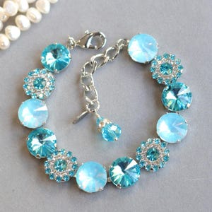 May include: A silver bracelet with light blue and turquoise crystal stones. The bracelet has round and flower-shaped crystals, linked by a chain and clasp. The background shows a strand of pearls. The bracelet's length is adjustable.