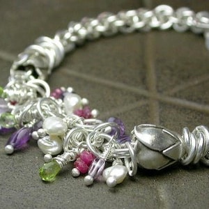 May include: A silver chain bracelet featuring a cluster of amethyst, ruby, and peridot gemstones. The bracelet has a decorative silver charm in the shape of a flower.