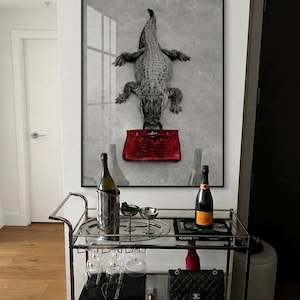 May include: A rectangular wall art print featuring a black and white crocodile holding a vibrant red handbag in its mouth. The artwork is displayed above a silver metal bar cart containing wine bottles, glassware, and a black quilted handbag.