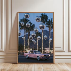 以下が含まれることがあります： ピンクのビンテージカーが、ディスコボールで飾られたヤシの木が並ぶ通りを走っています。車は、クロームバンパーと長いボンネットを持つ、クラシックなアメリカの筋肉質な車です。