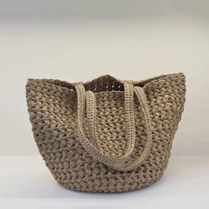 Può includere: Una borsa tote beige realizzata con un motivo intrecciato a maglia aperta. La borsa presenta due robusti manici intrecciati abbinati fissati al bordo superiore, creando una silhouette strutturata e flessibile con una texture in fibra naturale.