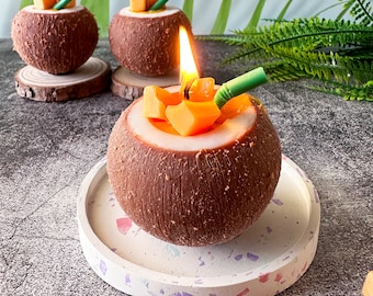 May include: A brown coconut shell candle with a lit wick and a yellow and orange fruit candle inside. The candle is sitting on a white and gray speckled round coaster.